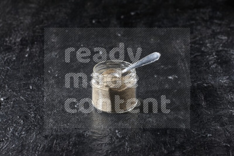 A glass jar and a metal spoon full of white pepper powder on textured black flooring