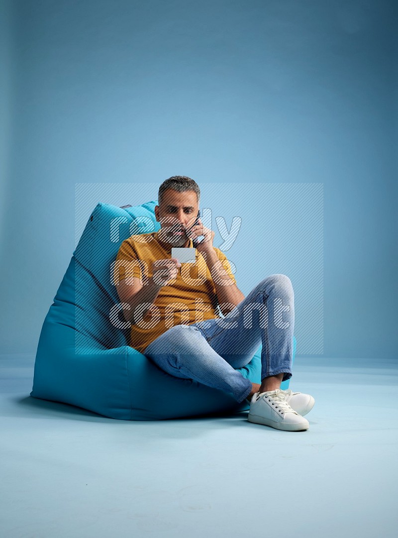 A man sitting on a blue beanbag and holding ATM card with phone