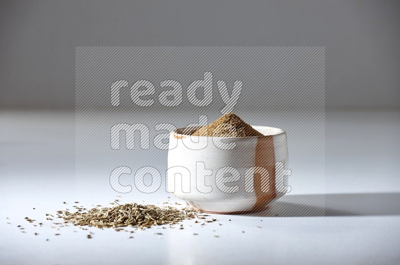 A beige bowl full of cumin powder with cumin seeds beneath it the bowl on a white flooring