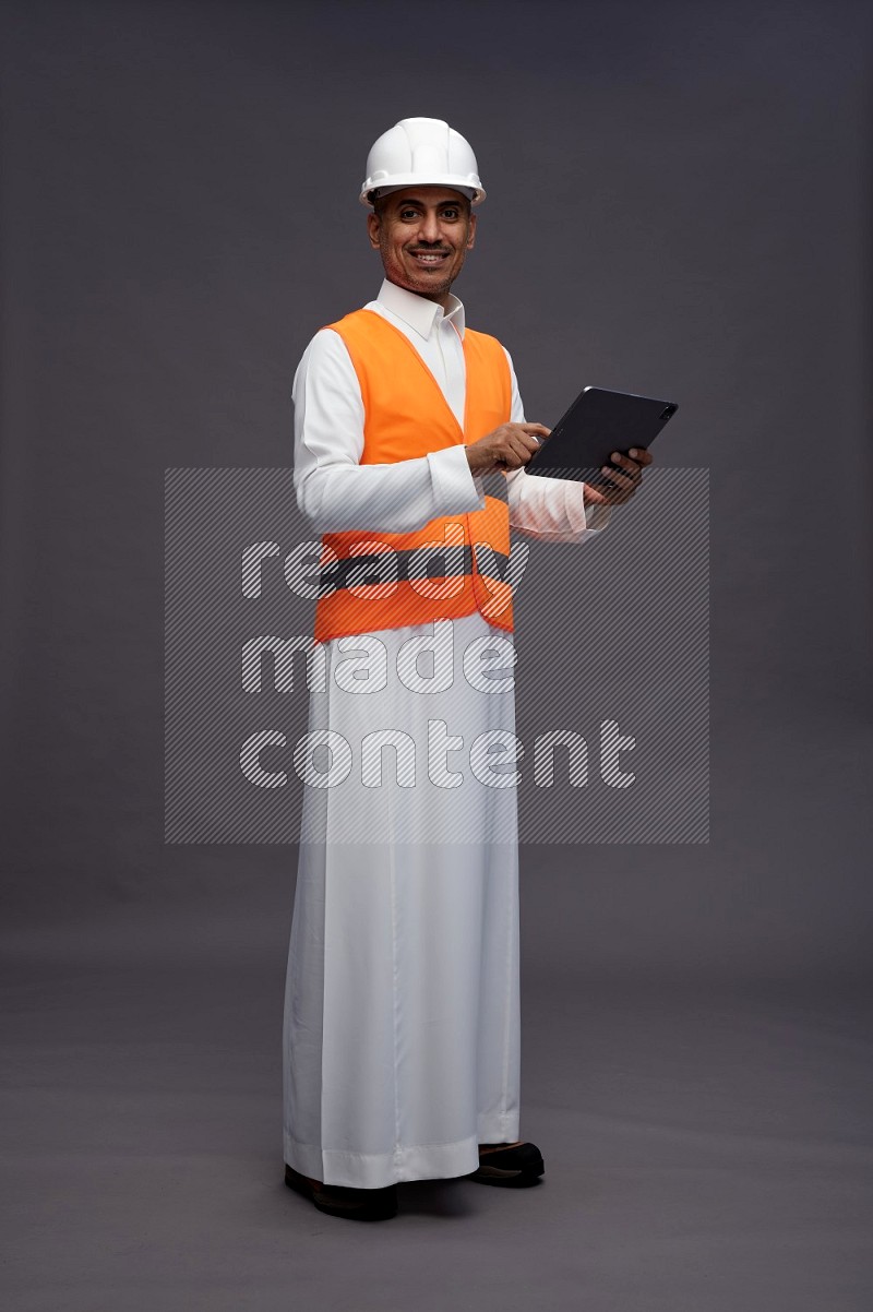 Saudi man wearing thob with engineer vest standing working on tablet on gray background