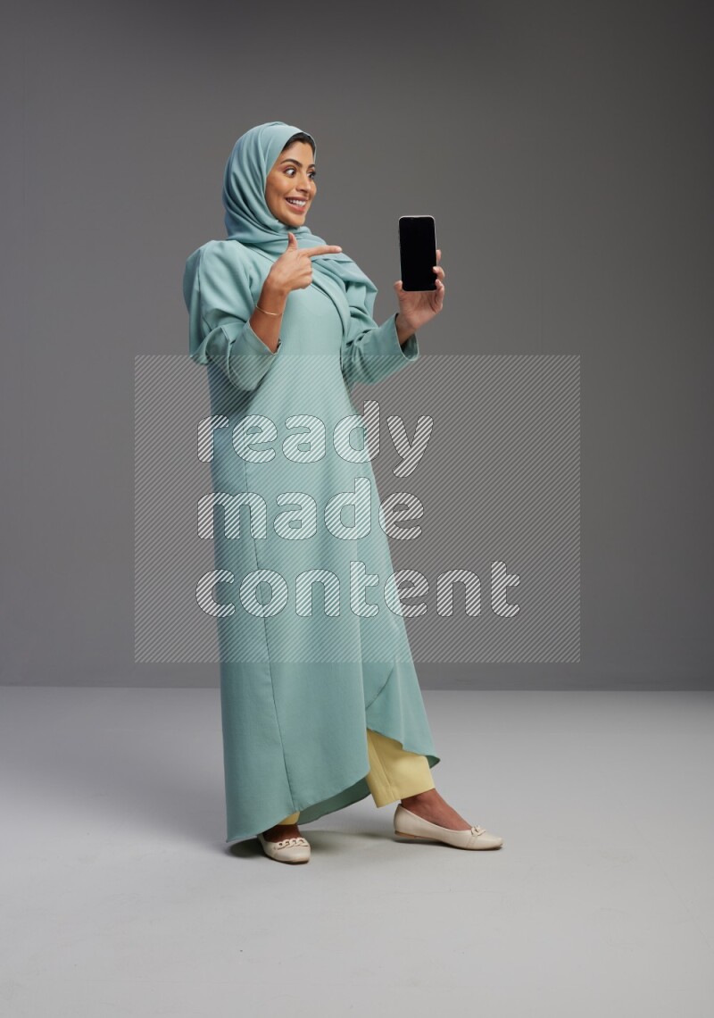 Saudi Woman wearing Abaya standing showing phone to camera on Gray background