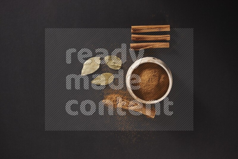 Cinnamon powder in a white pottery bowl and cinnamon sticks and laurel leaves on black background