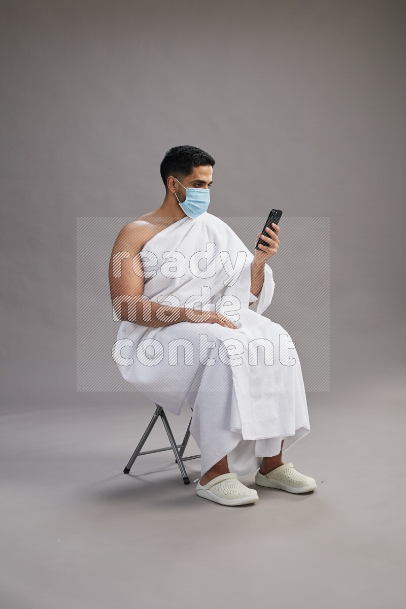 A man wearing Ehram with face mask sitting on chair texting on phone on gray background