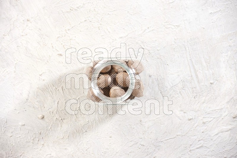 A glass spice jar full of nutmeg on a textured white flooring in different angles