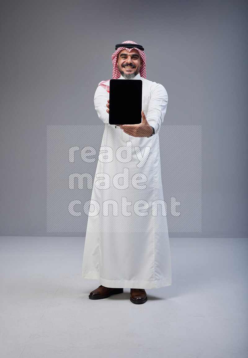 Saudi man Wearing Thob and red Shomag standing showing tablet to camera on Gray background