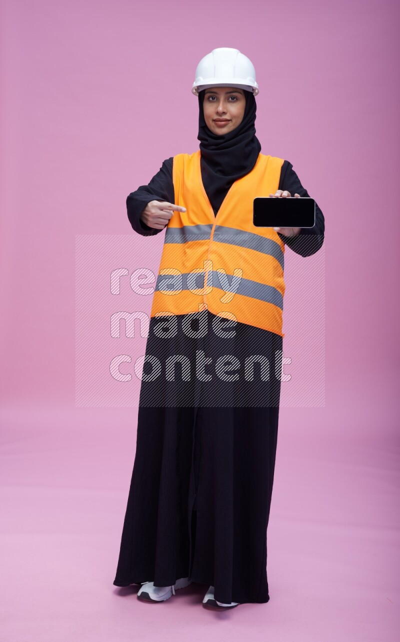 Saudi woman wearing Abaya with engineer vest and helmet standing showing phone to camera on pink background