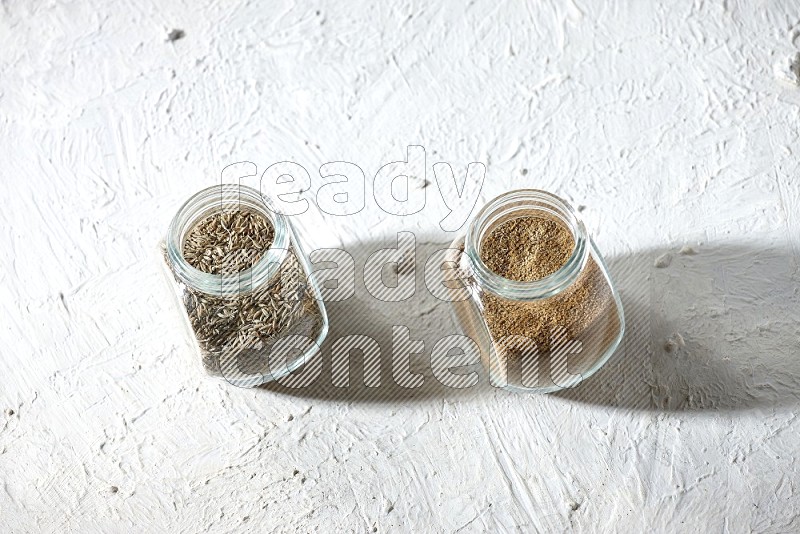 2 glass spice jars full of cumin powder and cumin seeds on textured white flooring