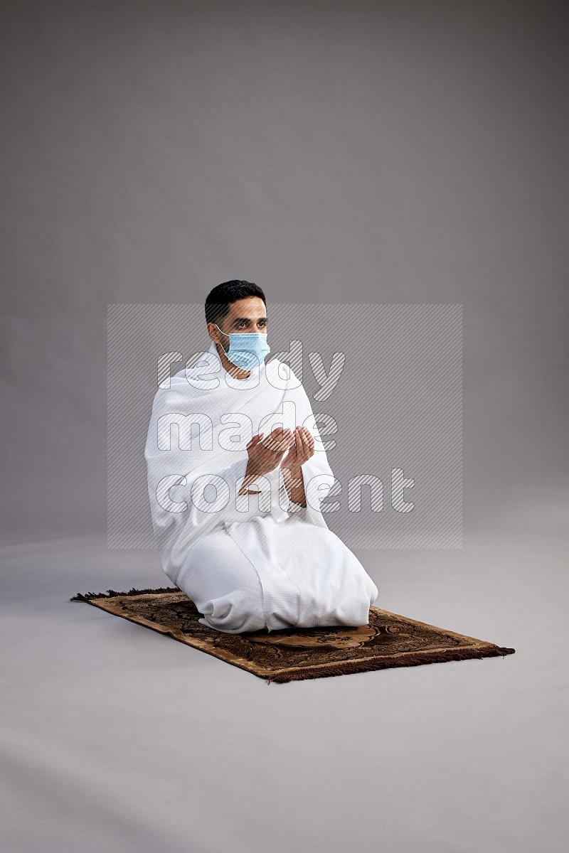 A man wearing Ehram with face mask sitting on floor performing dua'a on gray background