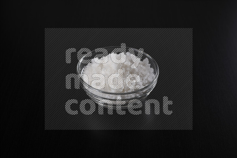 A glass bowl full of coarse sea salt crystals on black background