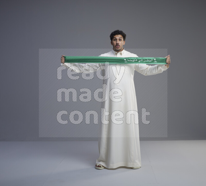 A Saudi man standing wearing thob holding Saudi flag scarf on gray background