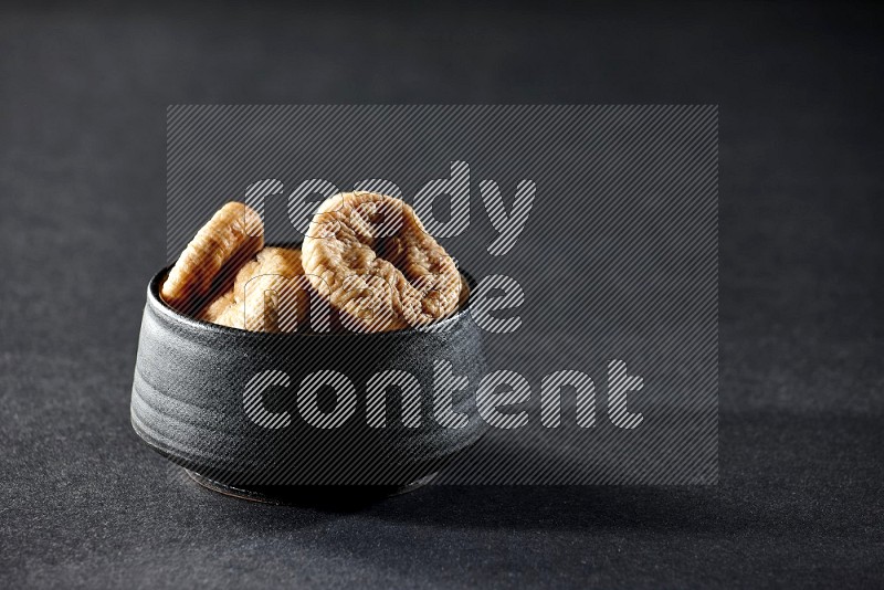 A black pottery bowl full of dried figs on a black background in different angles