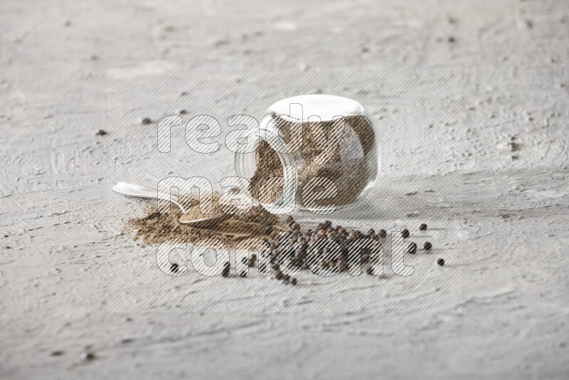 Flipped glass spice jar full of black pepper powder with a metal spoon and black pepper beads spread on a textured white flooring