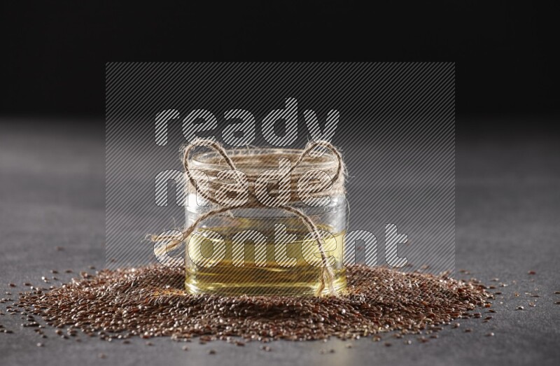 A glass jar full of flaxseeds oil surrounded by the seeds on a black flooring