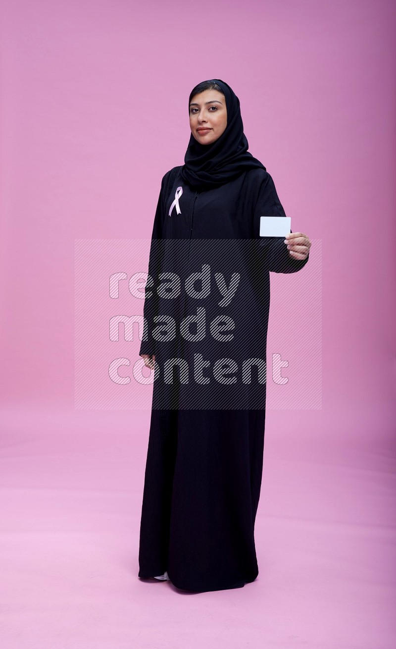Saudi woman wearing pink ribbon on Abaya standing holding ATM card on pink background