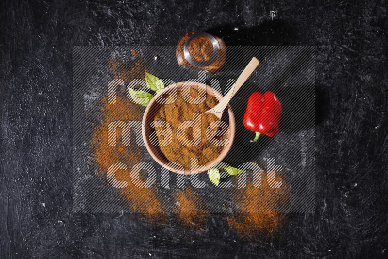 A wooden bowl full of ground paprika powder with a glass jar beside it and a red bell pepper on black background