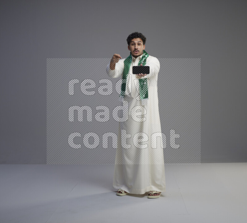 A Saudi man standing wearing thob and Saudi flag scarf showing phone to camera on gray background
