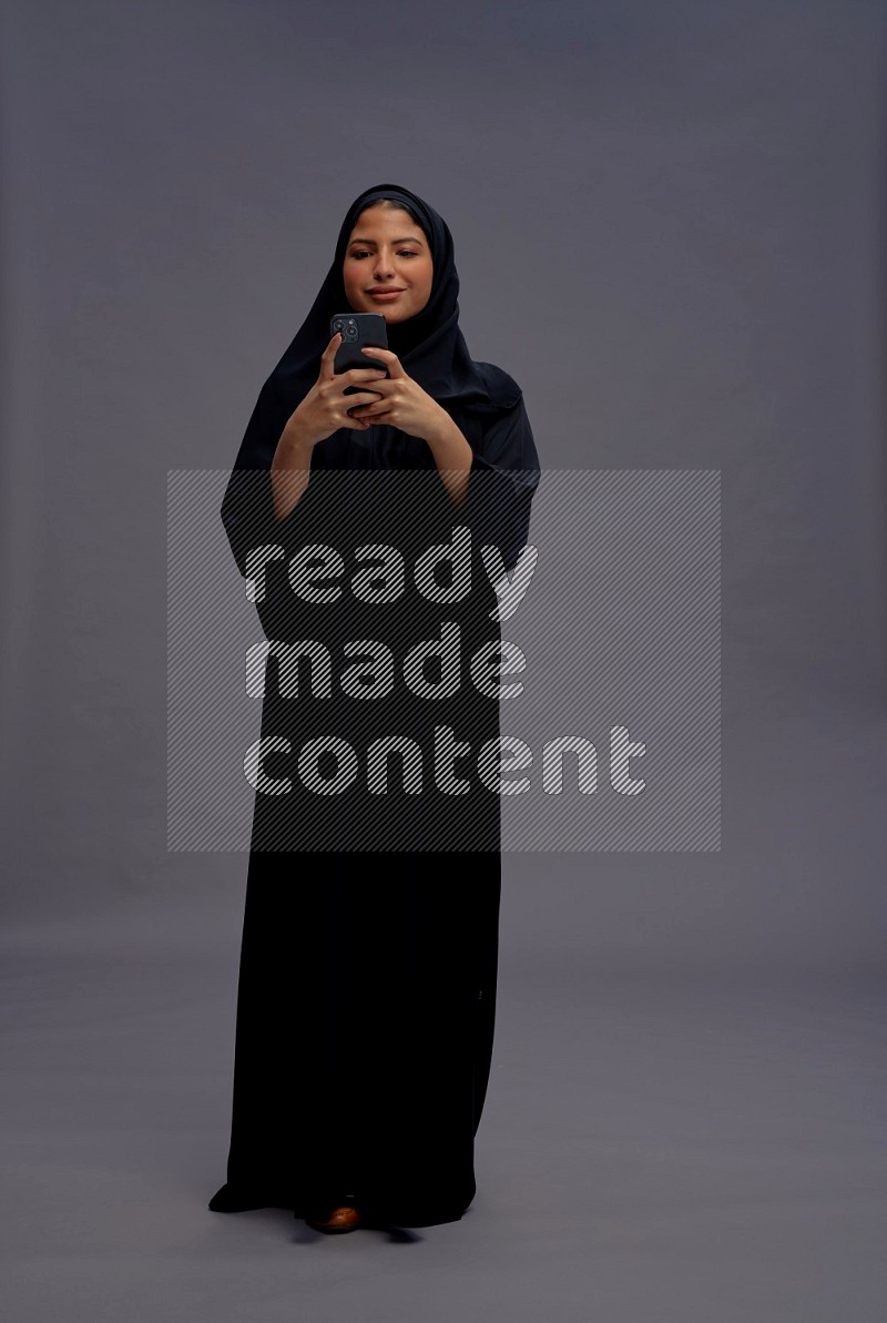 Saudi woman wearing Abaya standing texting on phone on gray background