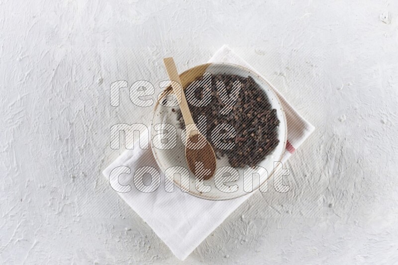 A Pottery plate full of whole cloves and a wooden spoon full of cloves powder in it on a textured white background
