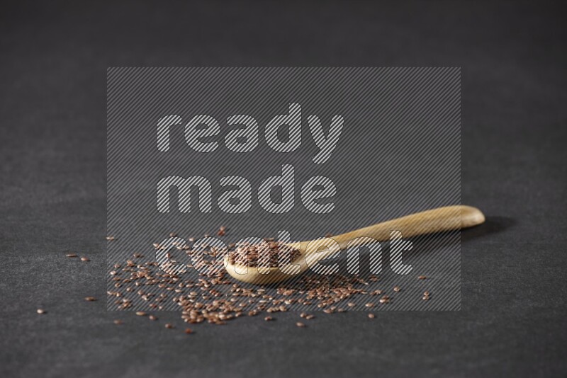 A wooden spoon full of flaxseeds and seeds spread beside it on a black flooring
