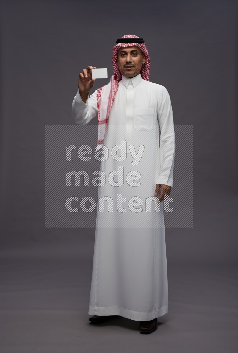 Saudi man wearing thob and shomag standing holding ATM card on gray background