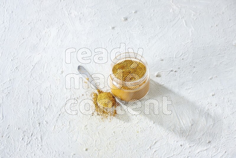 A glass jar and a metal spoon full of turmeric powder on a textured white flooring