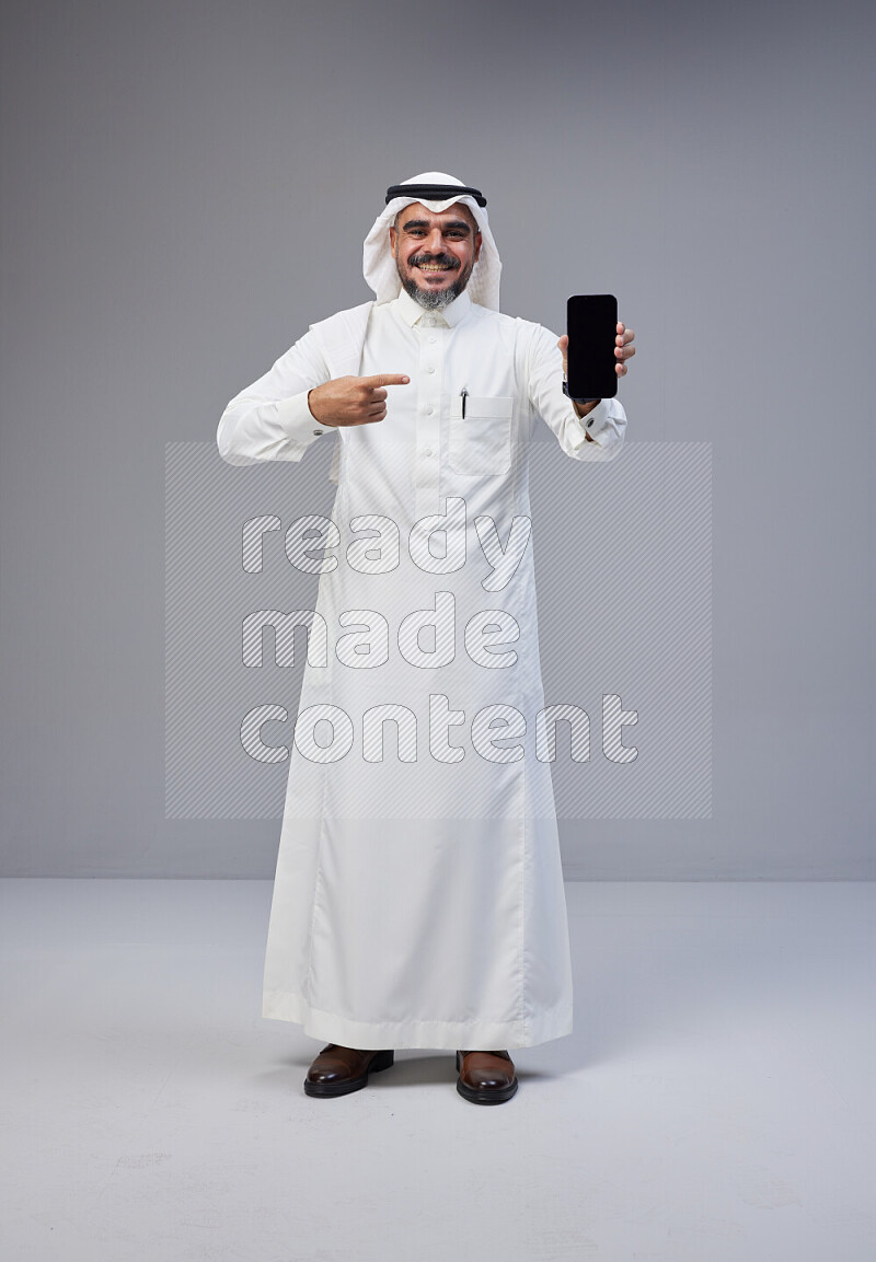 Saudi man Wearing Thob and white Shomag standing showing phone to camera on Gray background