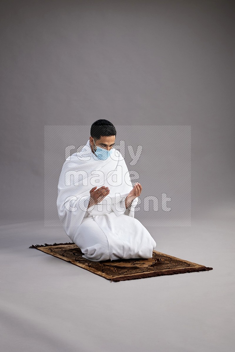 A man wearing Ehram with face mask sitting on floor performing dua'a on gray background