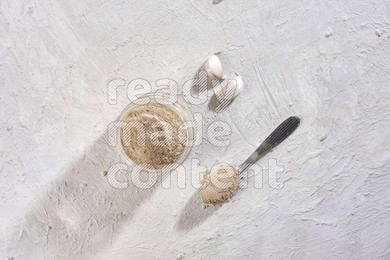 A glass jar full of garlic powder with metal spoon full of the powder on a textured white flooring in different angles