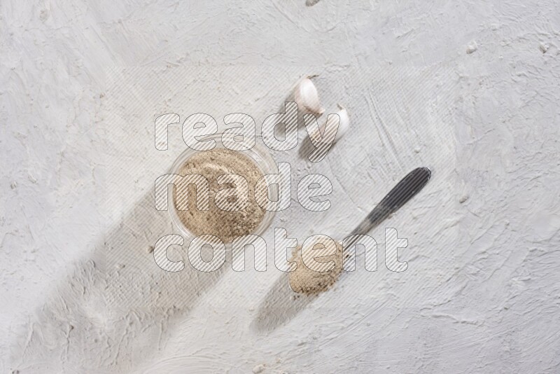 A glass jar full of garlic powder with a metal spoon full of the powder on a textured white flooring