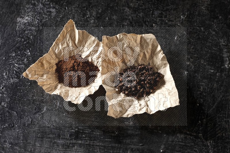 Cloves powder and cloves in 2 crumpled pieces of paper on a textured black flooring