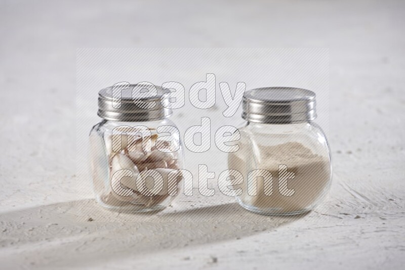 2 glass spice jars one full of garlic powder and the other full of cloves on a textured white flooring