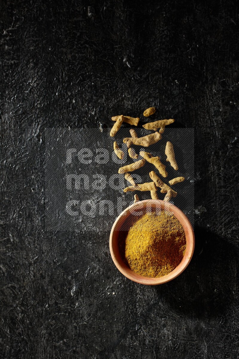 A wooden bowl full of turmeric powder with dried turmeric fingers on textured black flooring