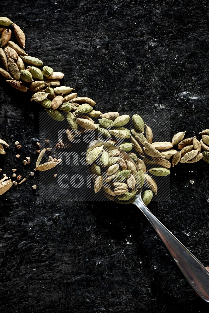 A Metal spoon full of cardamom seeds and some seeds beside it on a textured black flooring