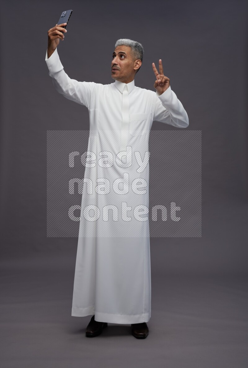 Saudi man wearing thob standing taking selfie on gray background