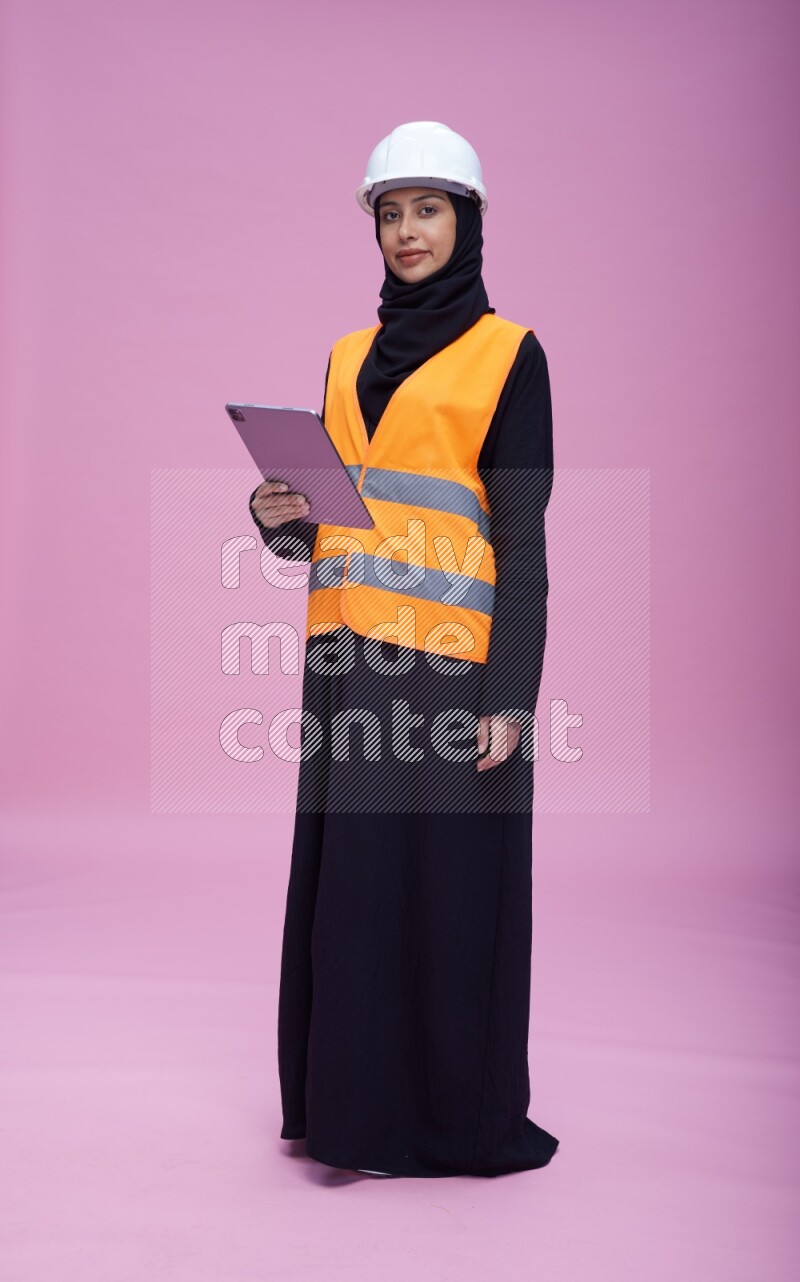 Saudi woman wearing Abaya with engineer vest and helmet standing working on tablet on pink background
