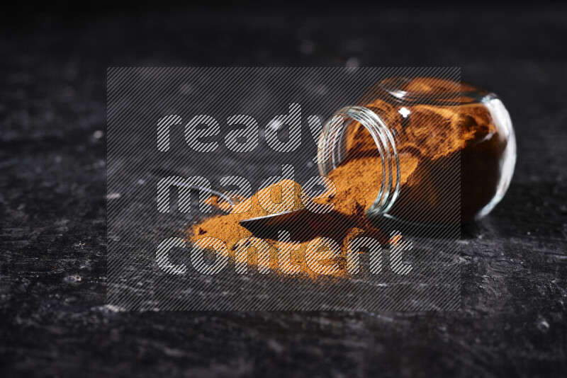 A glass jar full of ground paprika powder flipped with some spilling powder on black background