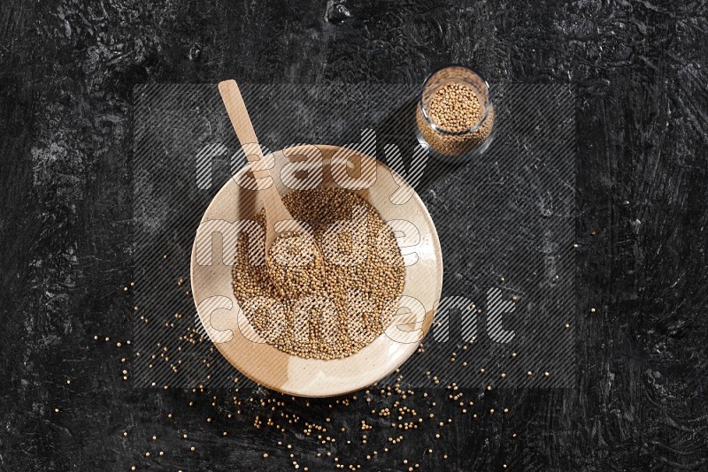 A multicolored pottery plate and a wooden spoon and a glass jar filled with mustard seeds on a textured black flooring in different angles