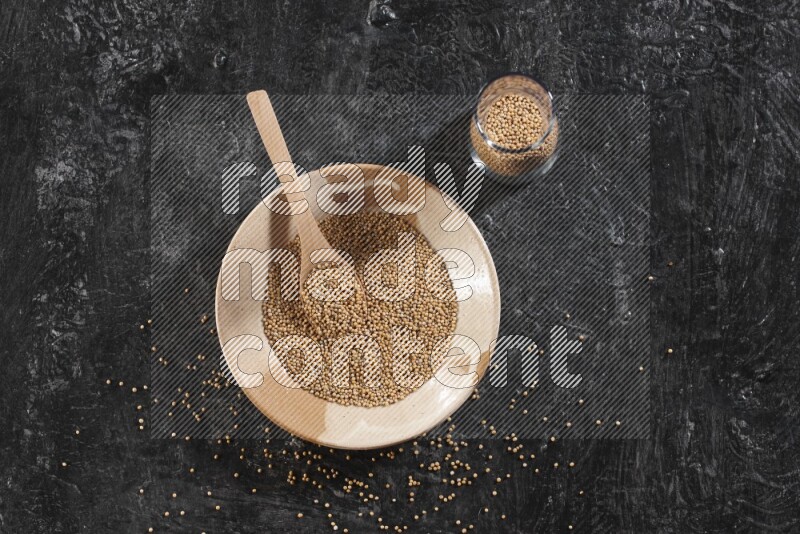 A beige pottery plate full of mustard seeds and a wooden spoon in it with a glass jar filled with the seeds on a textured black flooring