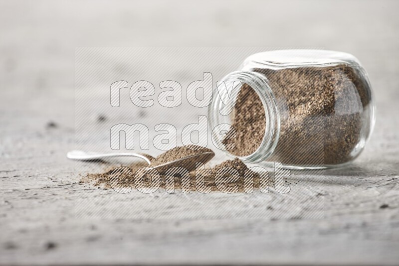 Flipped glass spice jar full of black pepper powder with a metal spoon full of it on textured white flooring