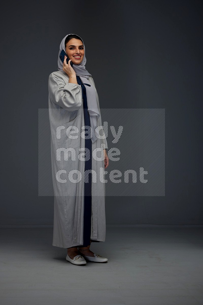 A Saudi woman wearing a light gray Abaya and head scarf standing and talking in the phone eye level on a grey background