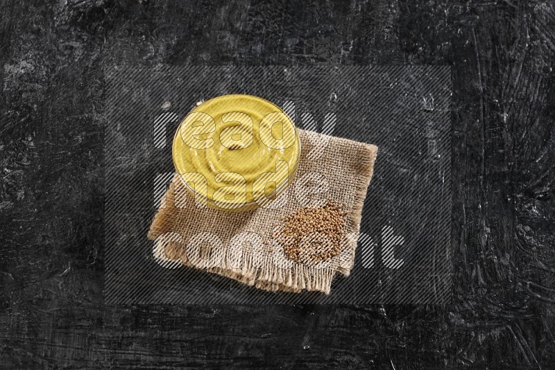 A glass bowl full of mustard paste set on a burlap piece with some mustard seeds on a textured black flooring
