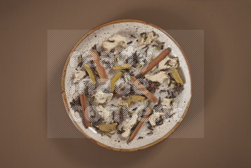 An assorted whole spices on a flat beige plate including fresh and dried ginger, cinnamon sticks, cardamom, turmeric fingers, cloves, star anise on a beige background