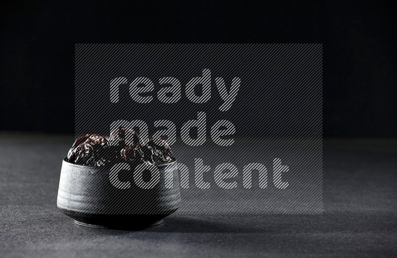 A black pottery bowl full of dried plums on a black background in different angles