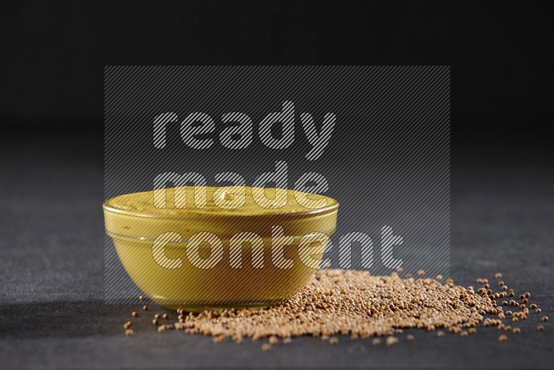 A glass bowl full of mustard paste with mustard seeds underneath on black flooring in different angles