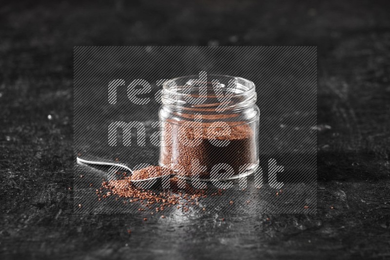 A glass jar full of garden cress seeds with a metal spoon full of the seeds on a textured black flooring