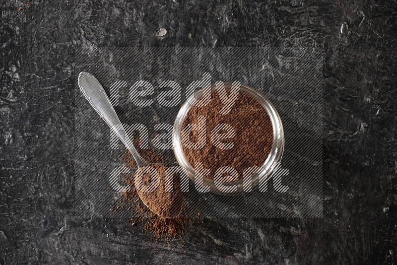 A glass jar full of cloves powder with a metal spoon on a textured black flooring