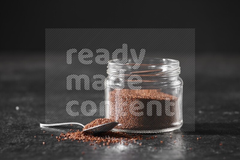 A glass jar full of garden cress seeds with a metal spoon full of the seeds on a textured black flooring