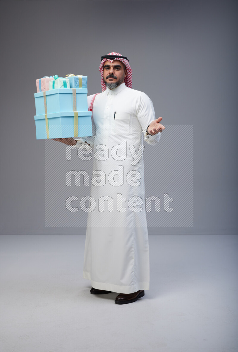 Saudi man Wearing Thob and red Shomag standing holding gift box on Gray background