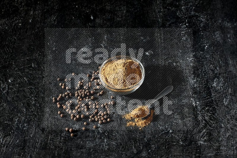 A glass bowl and metal spoon full of allspice powder and whole balls spreaded on a black flooring