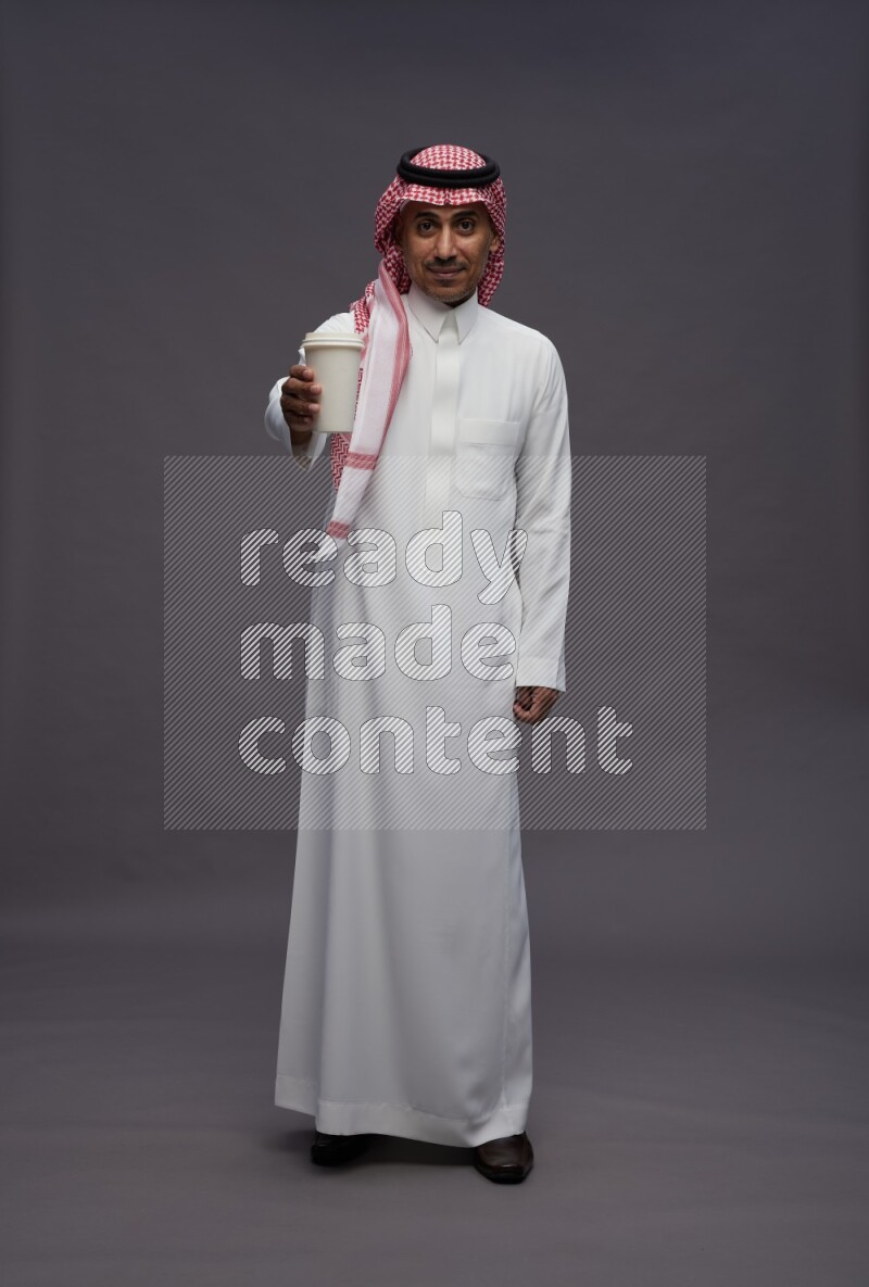 Saudi man wearing thob and shomag standing holding paper cup on gray background
